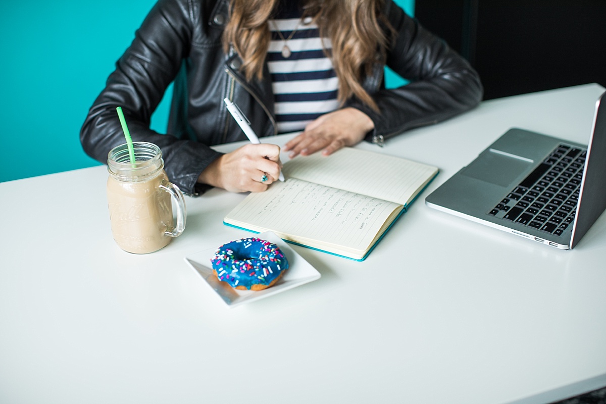 A woman writing in a journal with an iced coffee and blue frosted donut beside her