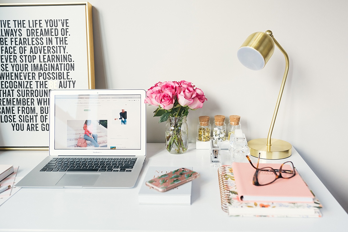 A pretty desk setup with a motivational poster, laptop, roses, pink notebook and gold lamp