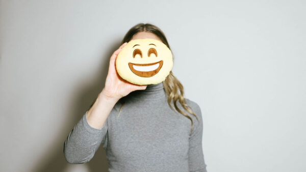 blonde woman wearing grey turtleneck holds a smiley face cookie in front of her face as part of a demo of emotional intelligence