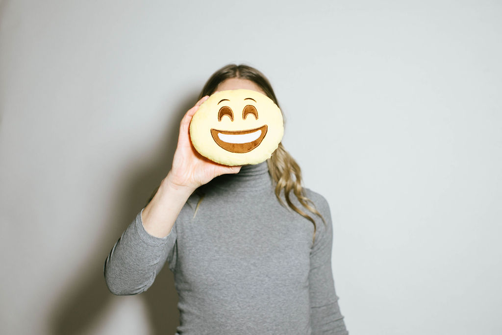 blonde woman wearing grey turtleneck holds a smiley face cookie in front of her face as part of a demo of emotional intelligence