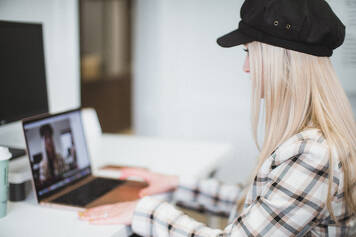blond woman wearing fancy plaid jacket and black brimmed hat sits at laptop talking to her team rocking remote workplace culture