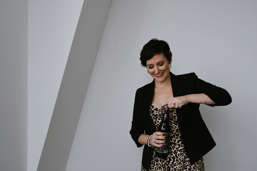 GIF of a beautiful woman with short, dark hair wearing a black blazer and leopard print dress cracking a beer and taking a drink in celebration of marketing services at meet pepper.