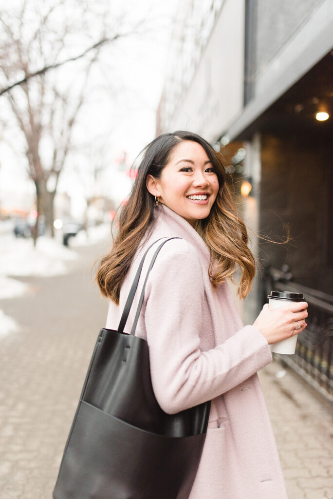 canadian asian woman wearing a pink jacket and holding a paper coffee cup looks back and smiles because she's excited about pepper's marketing service.