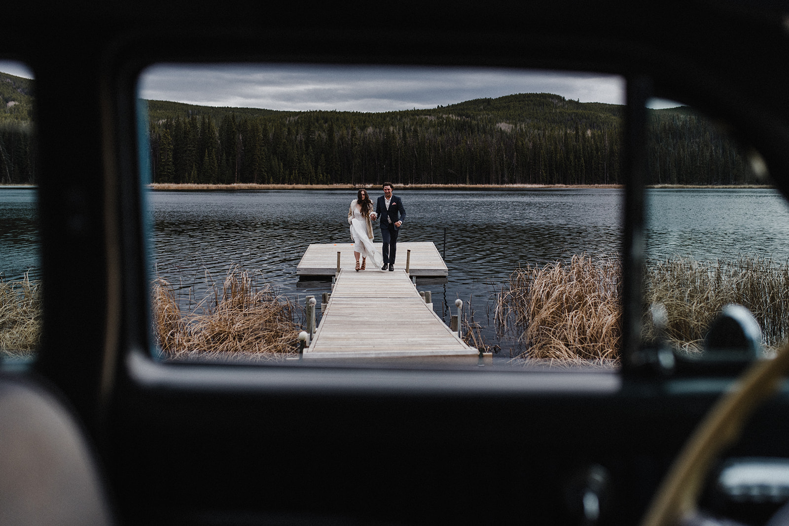 bride and groom framed on the end of a dock on a lake through an old-fashioned car window 