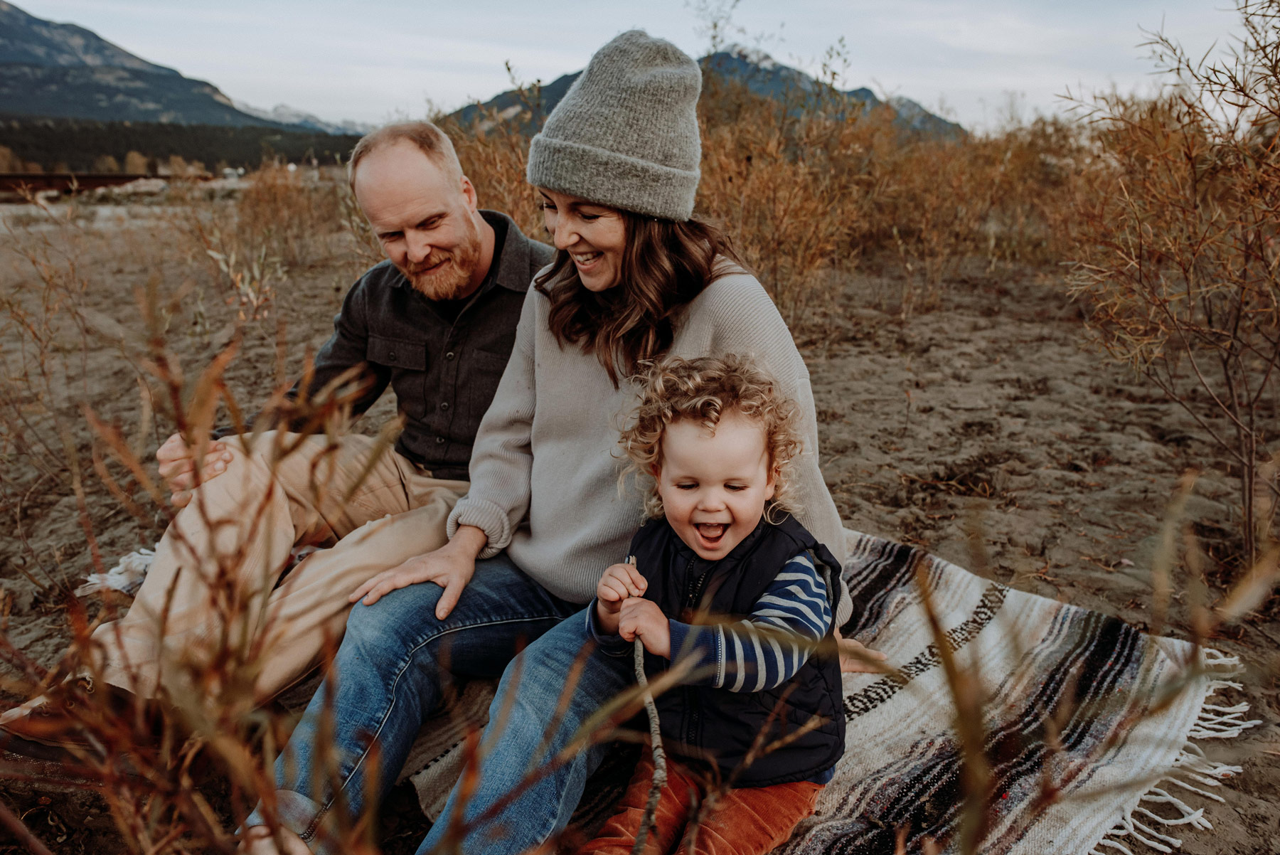 mom and dad with with curly haired little boy on a beach blanket laughing together in the fall in golden bc.