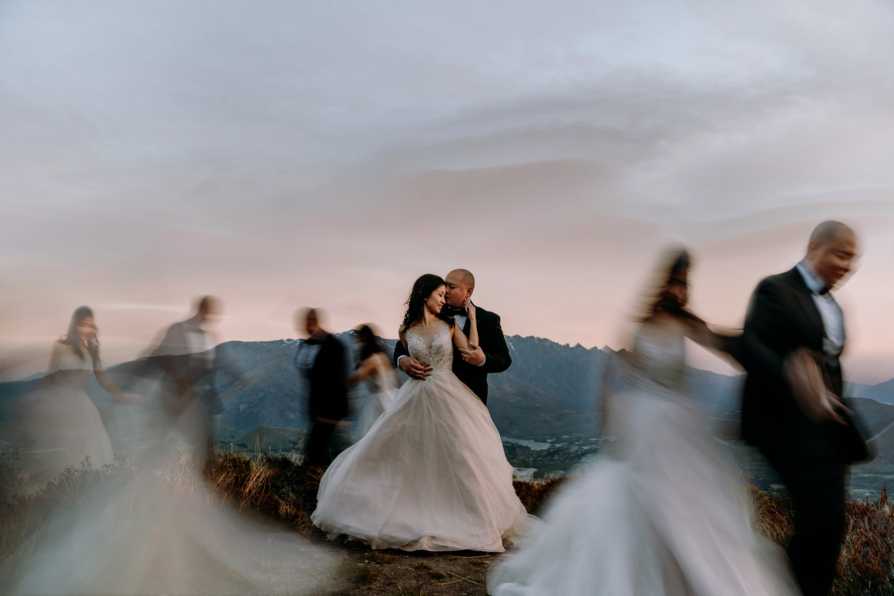 a bride and groom stand at the top of a mountain surrounding by blurred images of themselves walking circles arm in arm. 