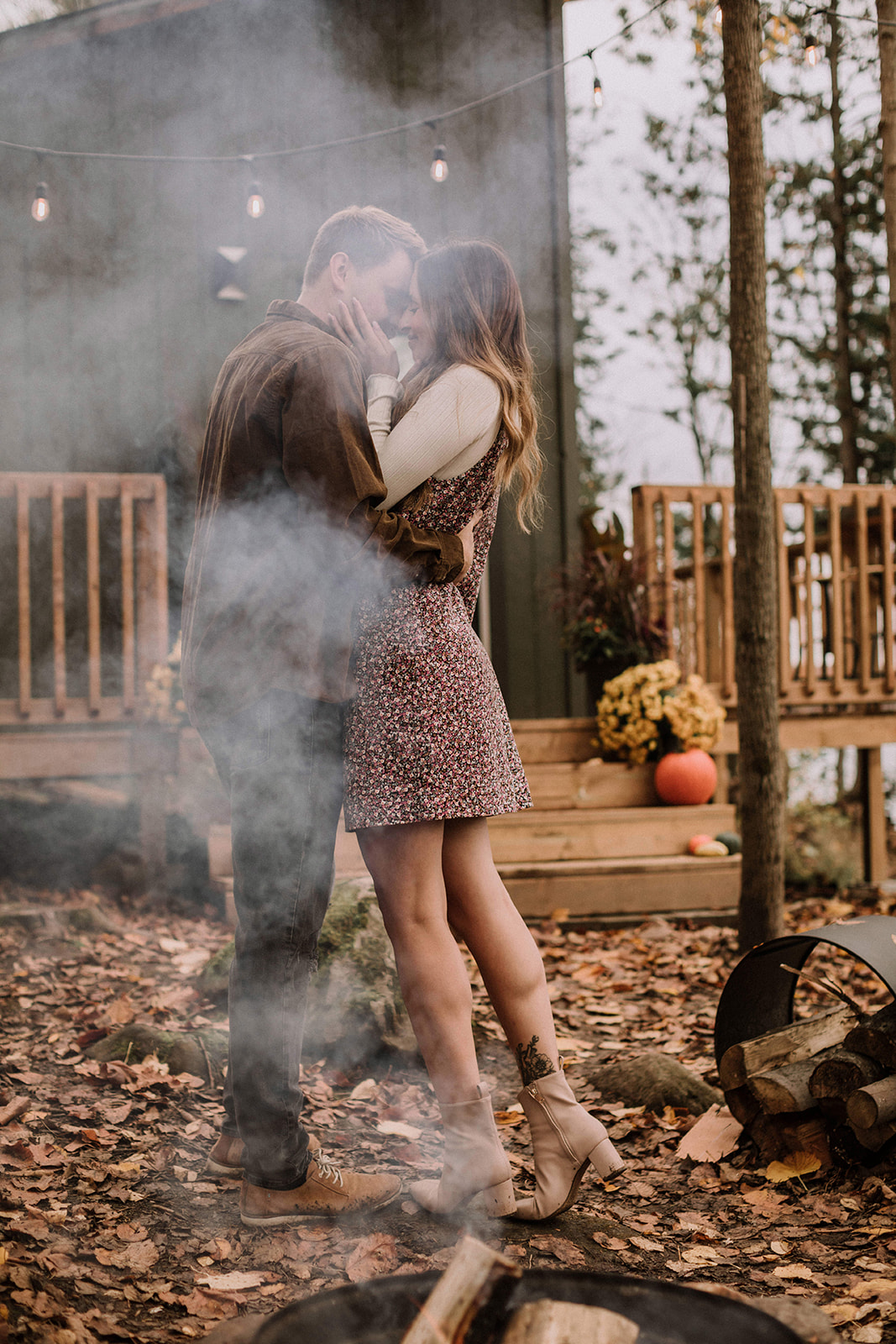 a young woman wearing a short floral print dress cuddles up to her fiance in front of their cabin at Lake Muskoka with a fire blazing in the first pit in front of them. 