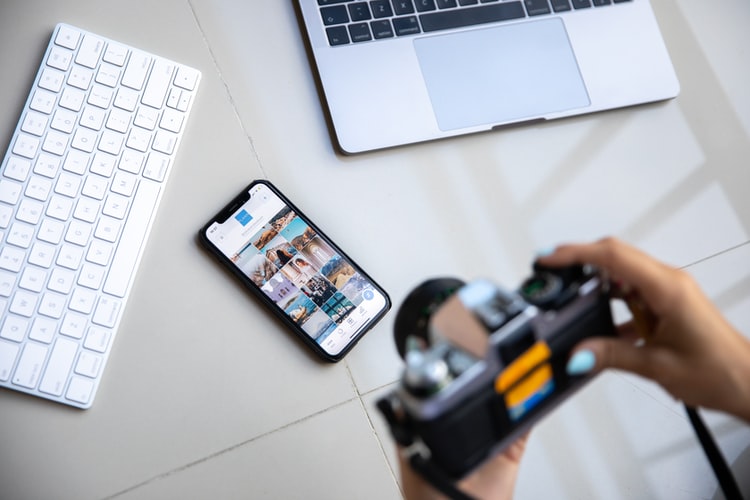 A photographer sitting at a desk with their camera, phone, and laptop, getting ready to make photo submissions as a public relations photography marketing tactic.