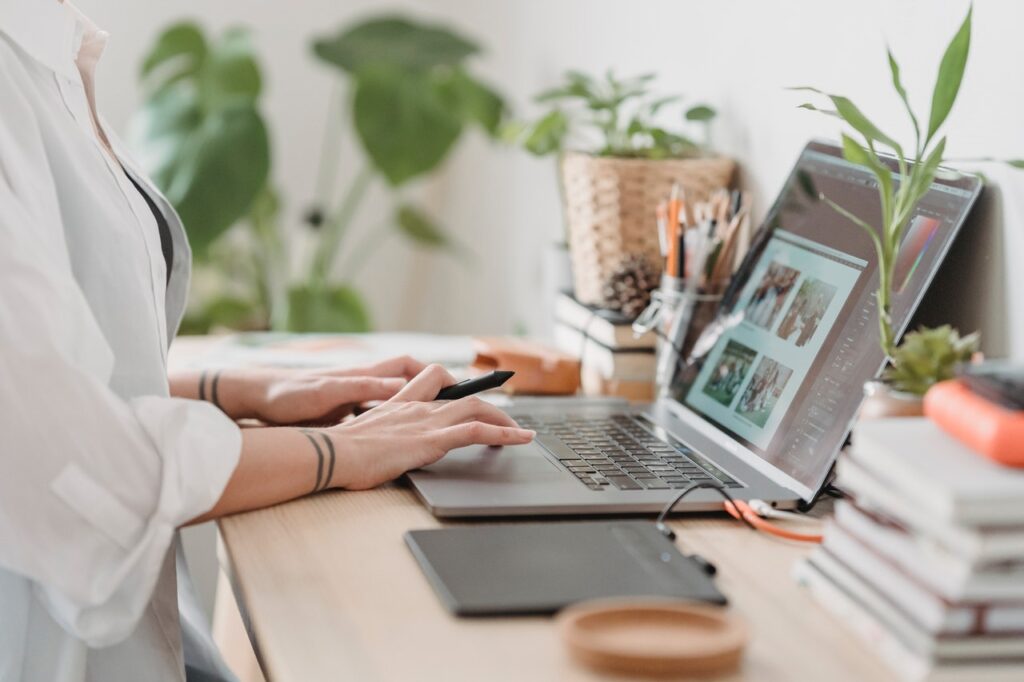 A tattooed photographer sitting at a desk working on a laptop, editing images to try to meet a photography deadline for a client.