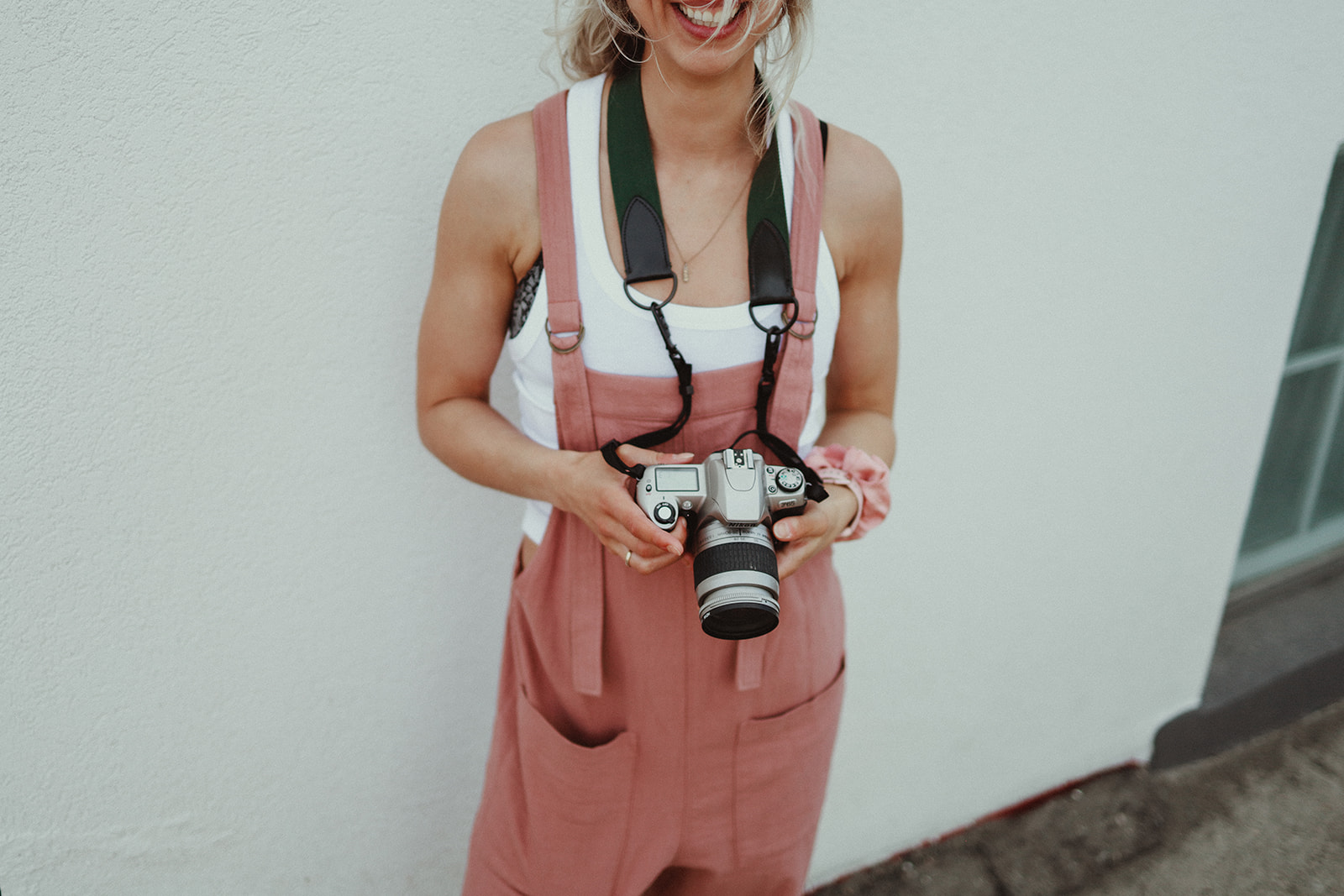 a blonde photographer wearing pink overalls holding a film camera and smiling because she has diversified marketing for her photography business.