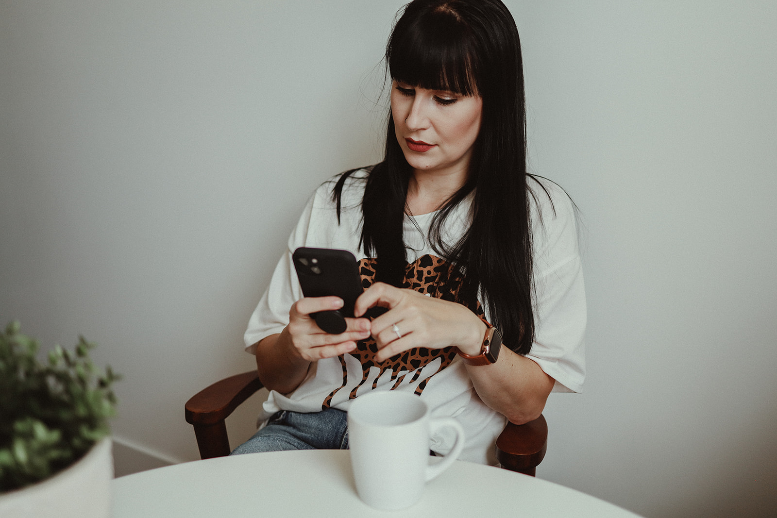 A photographer with long black hair and bangs sitting at a white table who is feeling the social media burnout scrolling on her phone while sipping a coffee trying to think of other ways to promote her photography business.
