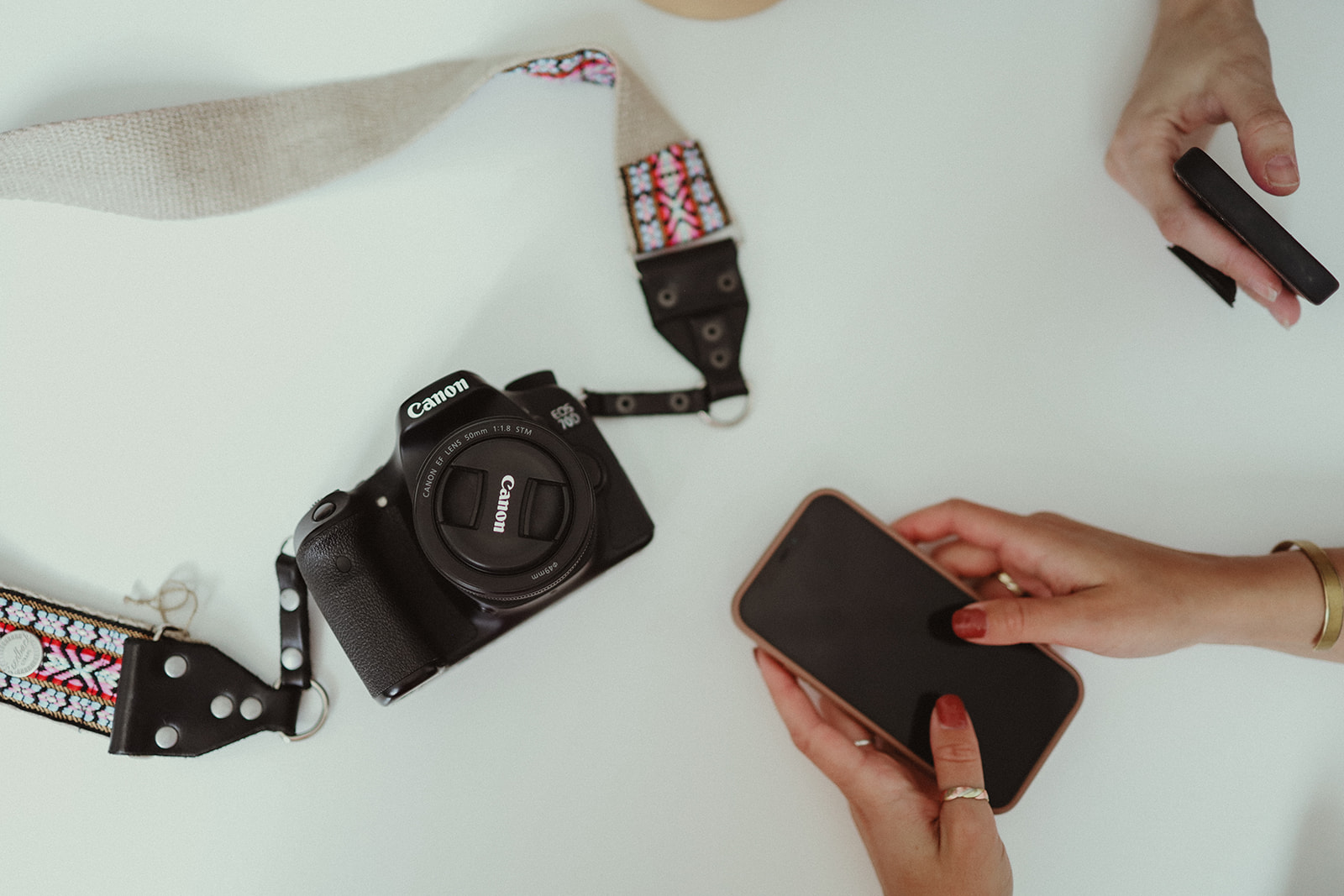 A canon camera with a pretty camera strap sitting on a white table beside two sets of hands looking on their phones researching SEO for photographers and how to increase website traffic.