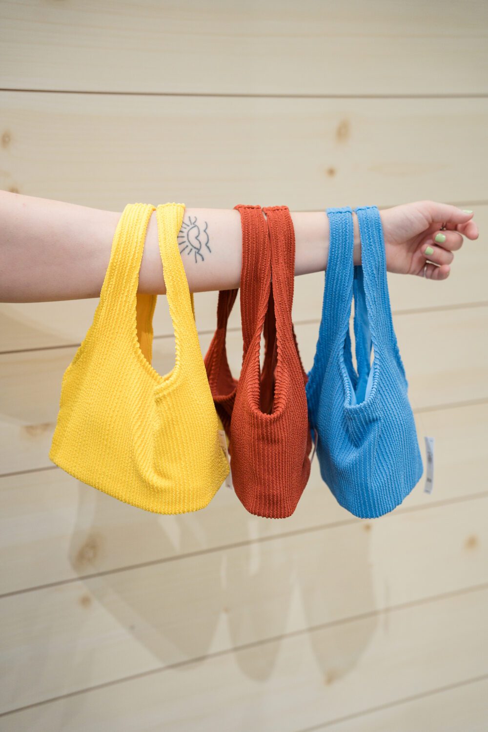 Three colourful textured handbags in yellow, rust, and blue hanging from someone’s arm inside a store at Aberdeen Mall.