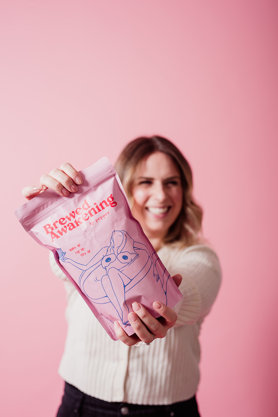 Woman holding a pink bag of Sweet Tooth Morning coffee toward the camera against a pink background during a branding photoshoot focused on product promotion and social media marketing.