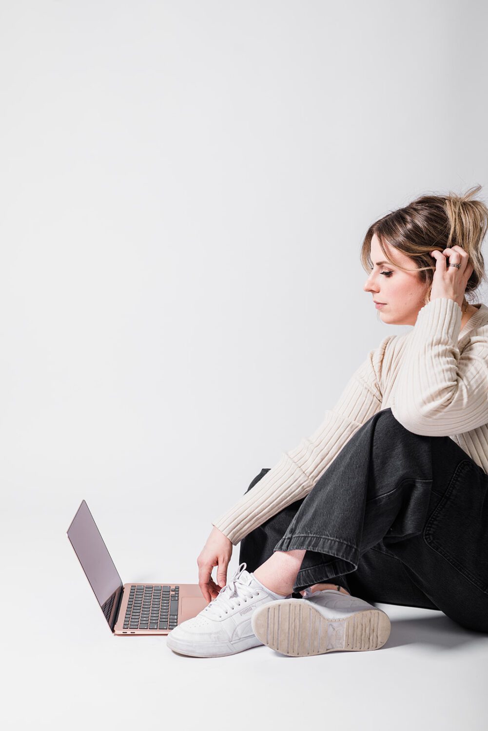 Woman sitting on a white studio floor using a laptop while working on marketing strategy and branding tasks, representing remote work and digital marketing agency services.