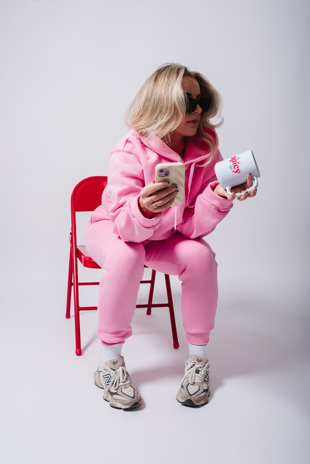 Woman in a pink tracksuit sitting on a red chair holding a coffee mug and coffee bag during a studio product shoot designed for social media branding and marketing content.
