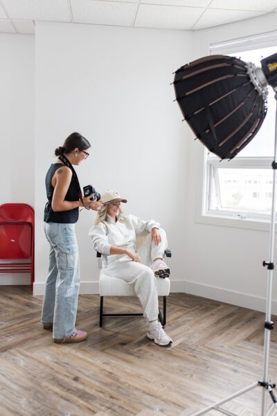 Behind-the-scenes photo of a marketing agency photoshoot in a bright studio, with a photographer adjusting lighting while a woman sits in a chair reviewing a laptop during content creation for branding and marketing materials.