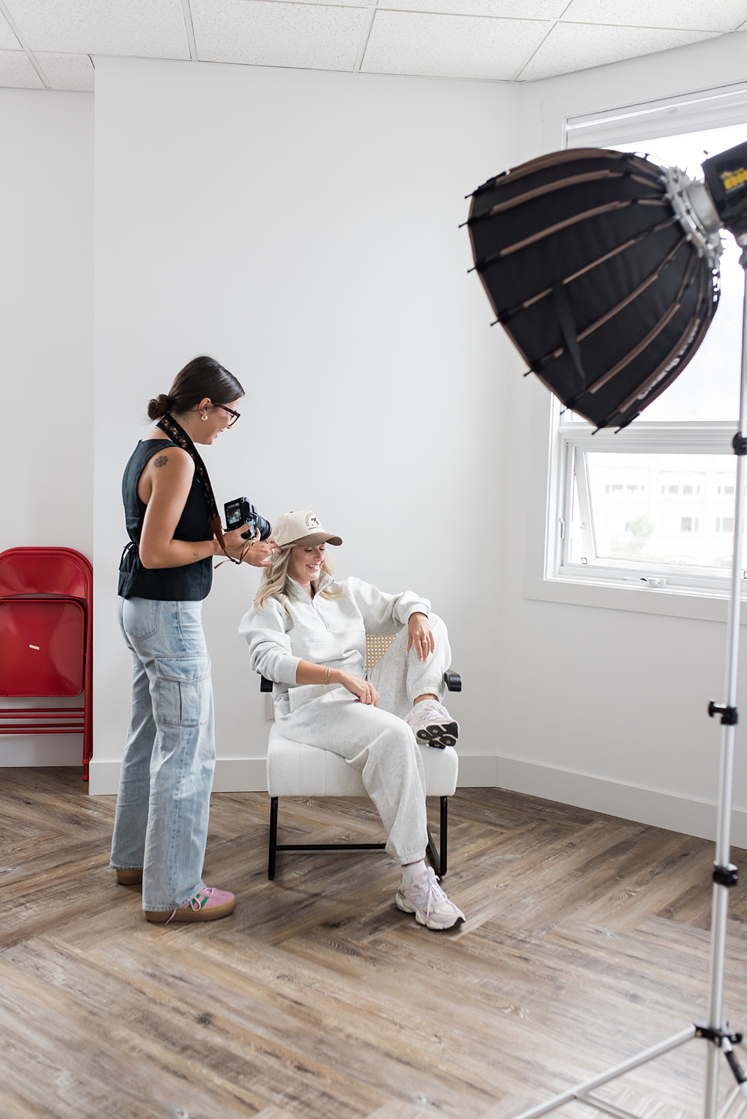 Behind-the-scenes photo of a marketing agency photoshoot in a bright studio, with a photographer adjusting lighting while a woman sits in a chair reviewing a laptop during content creation for branding and marketing materials.