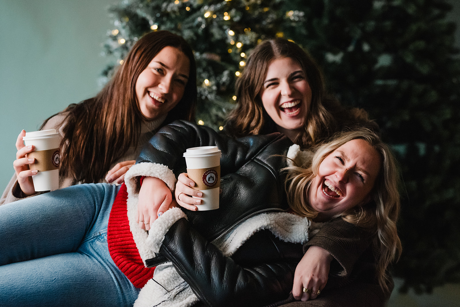 Three women laughing together while holding coffee in front of a decorated tree, capturing a warm, social winter moment in Kamloops for tourism and local lifestyle marketing.