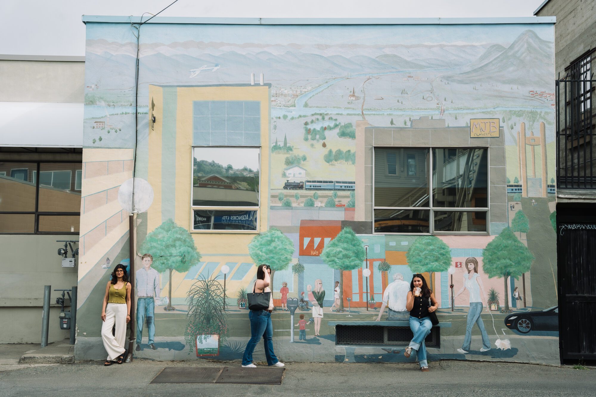 Three women pose and walk in front of a large downtown Kamloops mural showing buildings, mountains, trees, a train, and local street life.