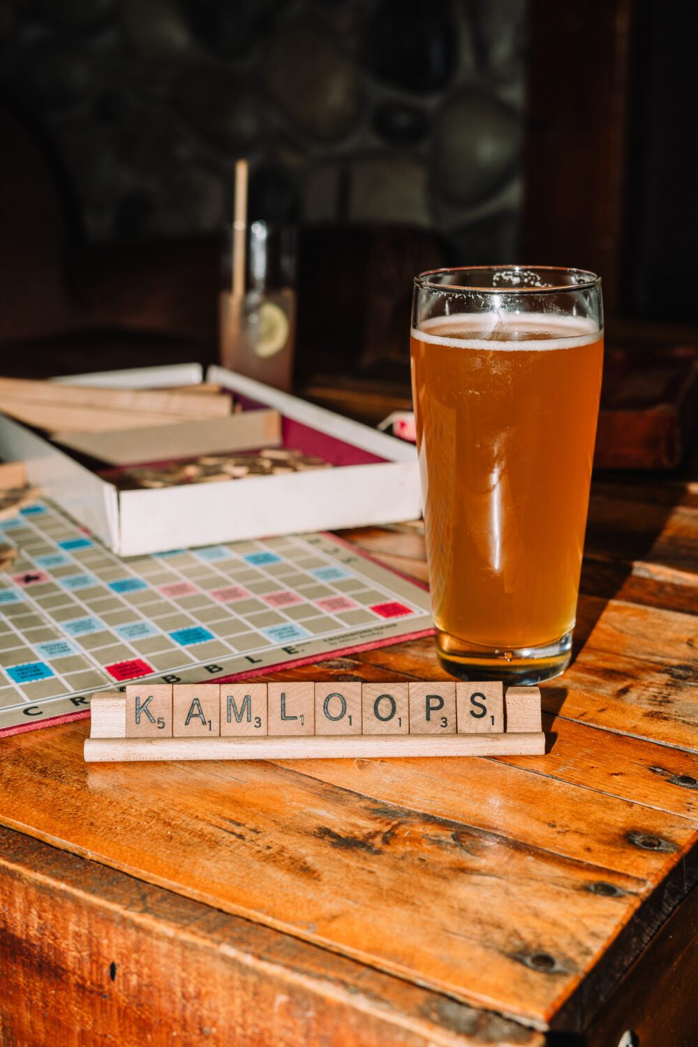 A pint of beer sits on a wooden table beside Scrabble tiles spelling “Kamloops” inside a downtown Kamloops brewery.
