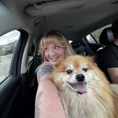 Woman smiling in a car while taking a selfie with a fluffy dog sitting beside her, capturing candid lifestyle content.