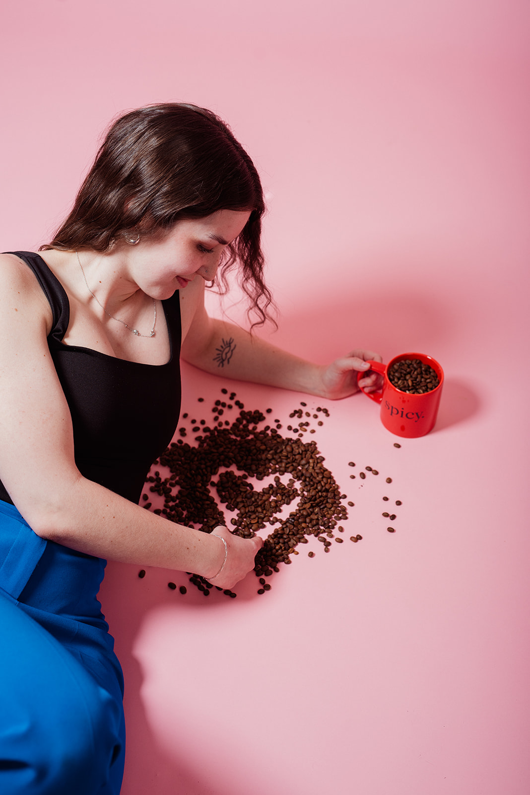 Woman lying on a pink studio floor arranging coffee beans into a heart shape beside a red mug during a creative lifestyle product shoot for a coffee brand’s marketing campaign.