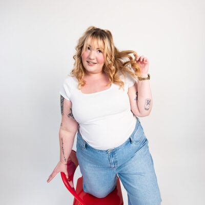 Woman leaning on a red chair wearing a white shirt and jeans during a casual studio portrait session for personal branding imagery.