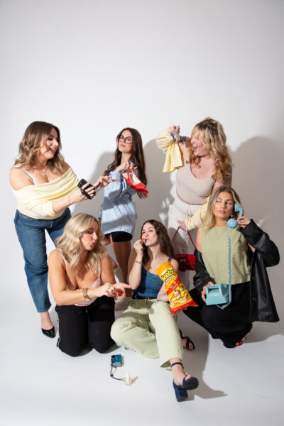 Group of women sitting and standing together in a studio while holding colorful accessories and handbags during a collaborative lifestyle branding photoshoot.