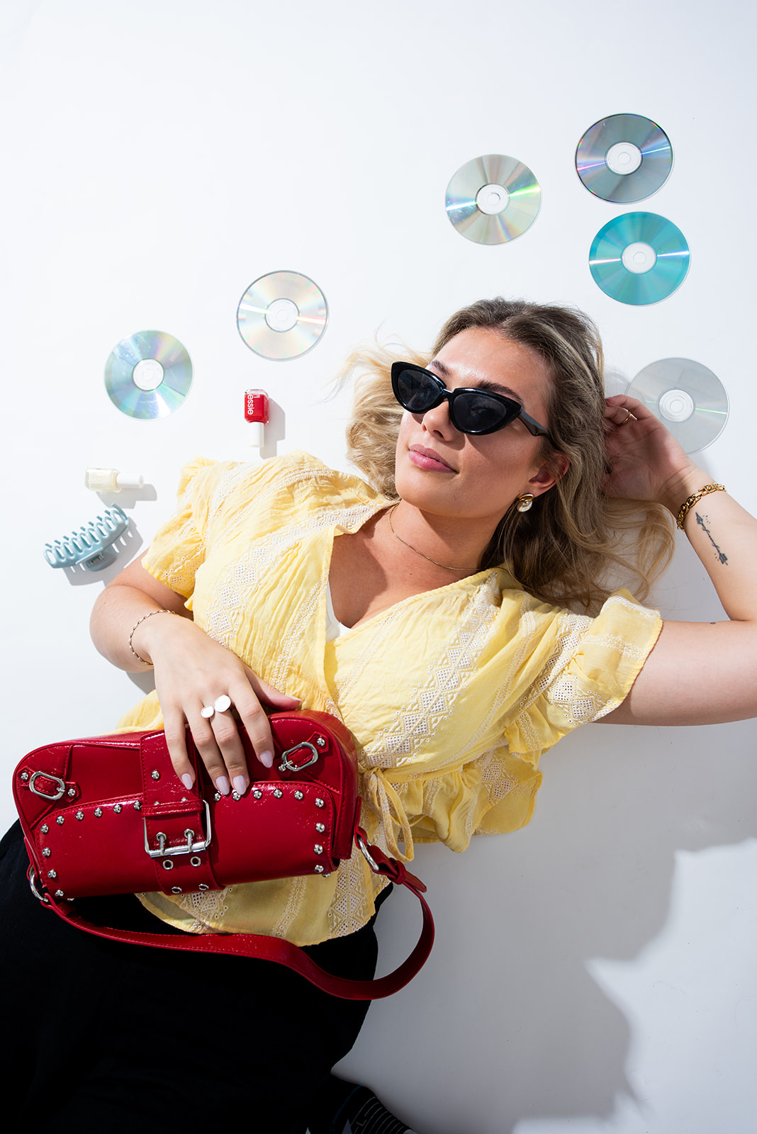 Woman in sunglasses reclining beside CDs, a keyboard, and a red purse in a bright studio for Pepper’s brand photography.