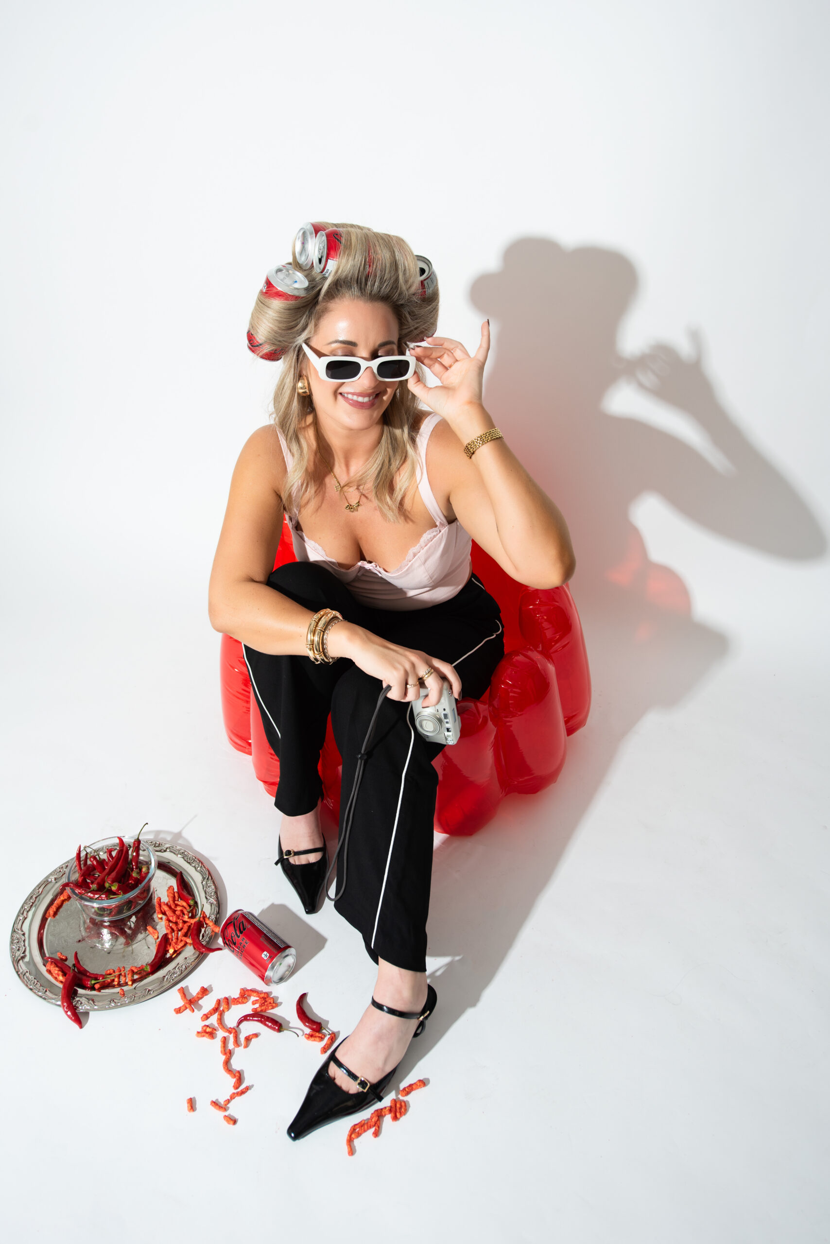 Woman sitting on a red inflatable chair wearing sunglasses and soda-can hair rollers during a playful lifestyle branding photoshoot designed for social media marketing content.