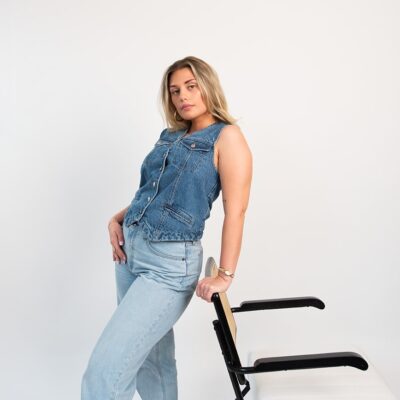 Woman leaning against a studio chair wearing a denim vest and light jeans during a fashion-inspired branding photoshoot.