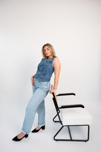 Woman leaning against a studio chair wearing a denim vest and light jeans during a fashion-inspired branding photoshoot.
