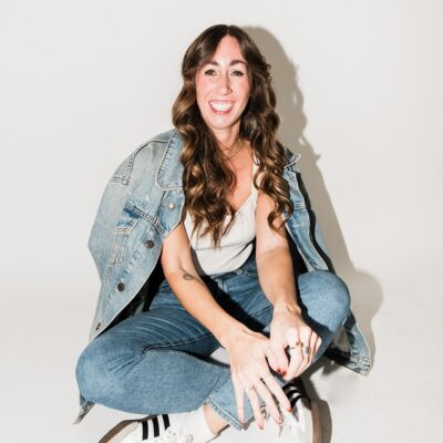 Woman sitting cross-legged wearing a denim jacket and jeans in a studio portrait used for personal branding photography.