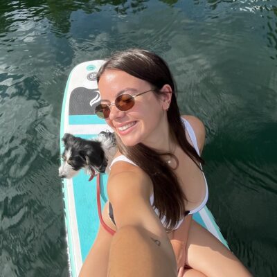 Woman paddleboarding on a lake with a small dog while taking a selfie, capturing outdoor lifestyle and adventure content.
