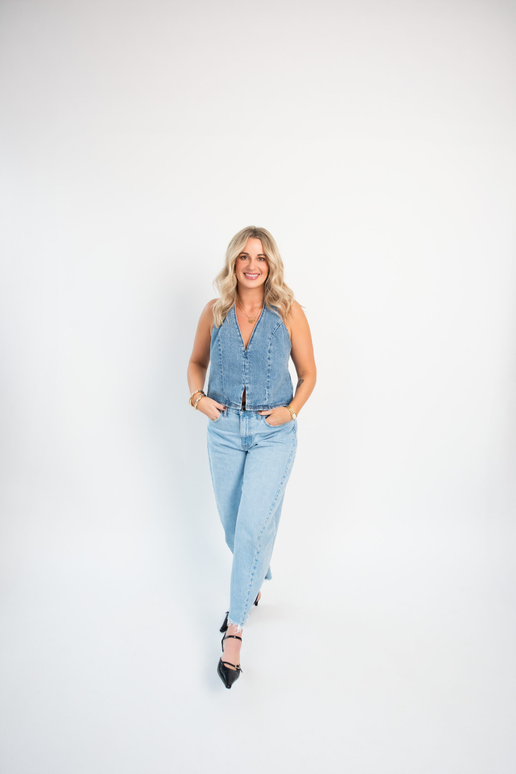 Blonde woman in a denim sleeveless top and jeans walking confidently toward camera against a clean white background, representing Pepper’s modern branding and marketing agency presence in Kamloops.
