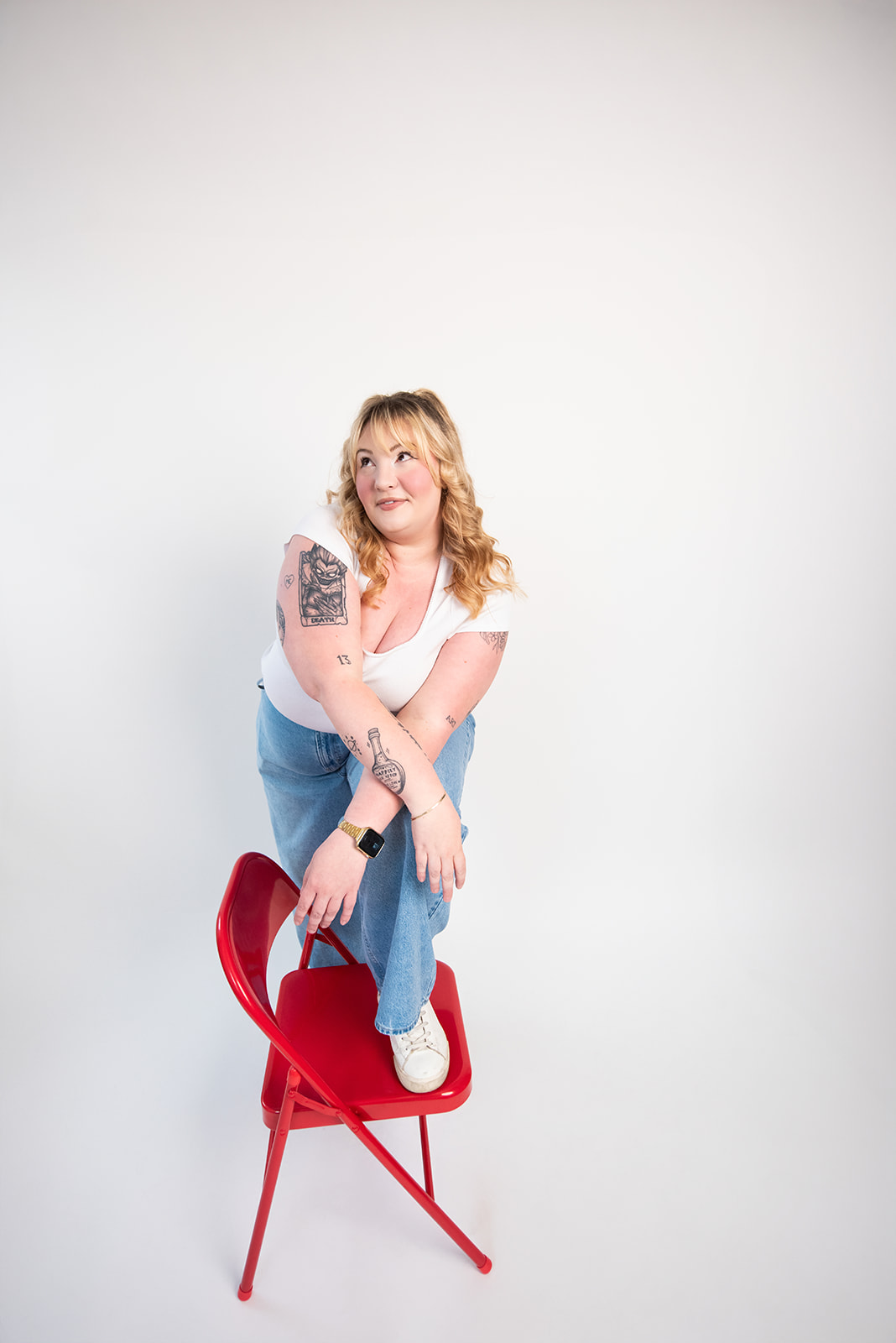 Woman in a white t-shirt and light denim overalls sitting on a red chair in a white studio, looking off to the side, reflecting Pepper’s creative, personality-led approach to branding and marketing in Kamloops.