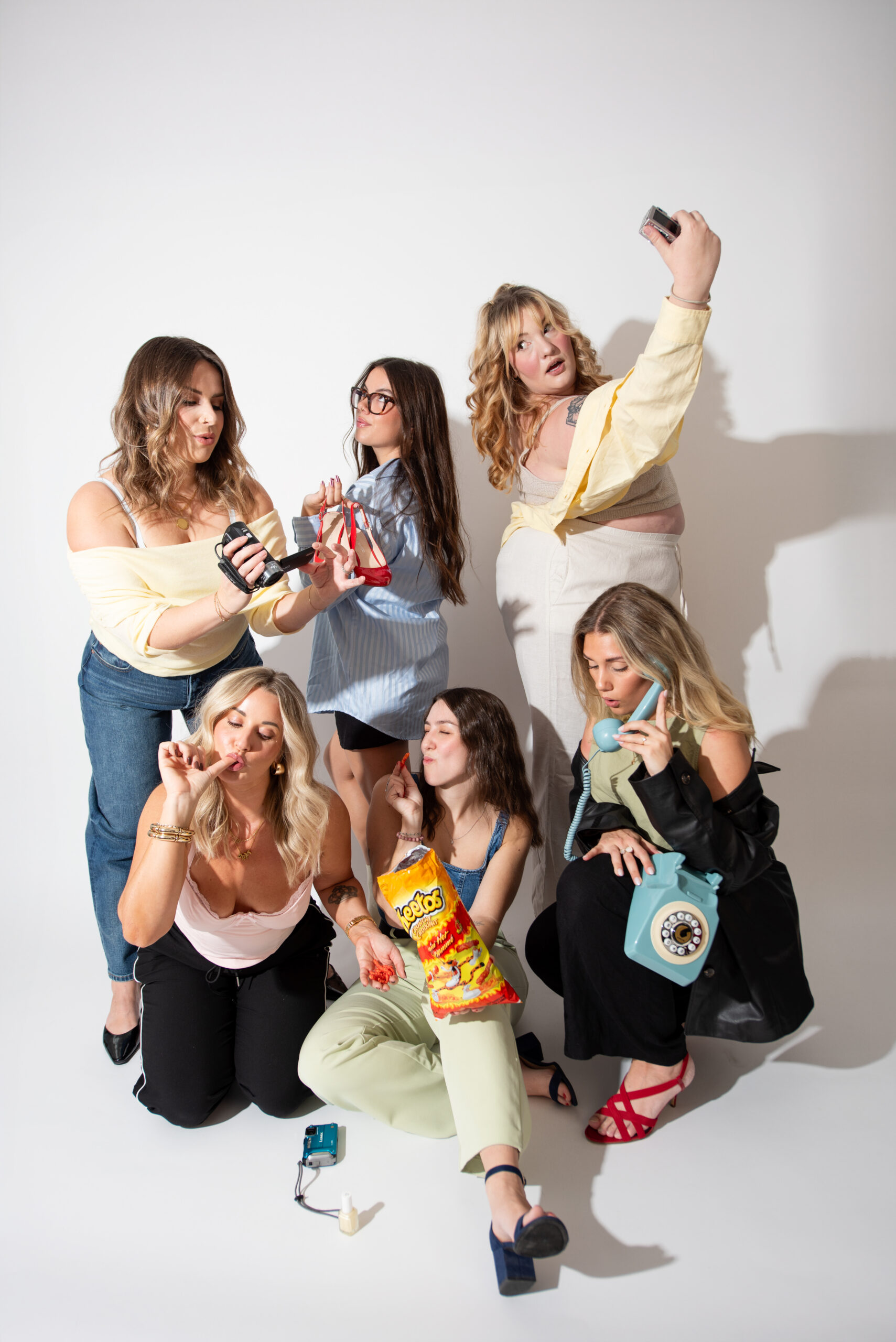 Six women posing together in a studio holding colorful handbags and accessories during a playful branding photoshoot for lifestyle marketing content.
