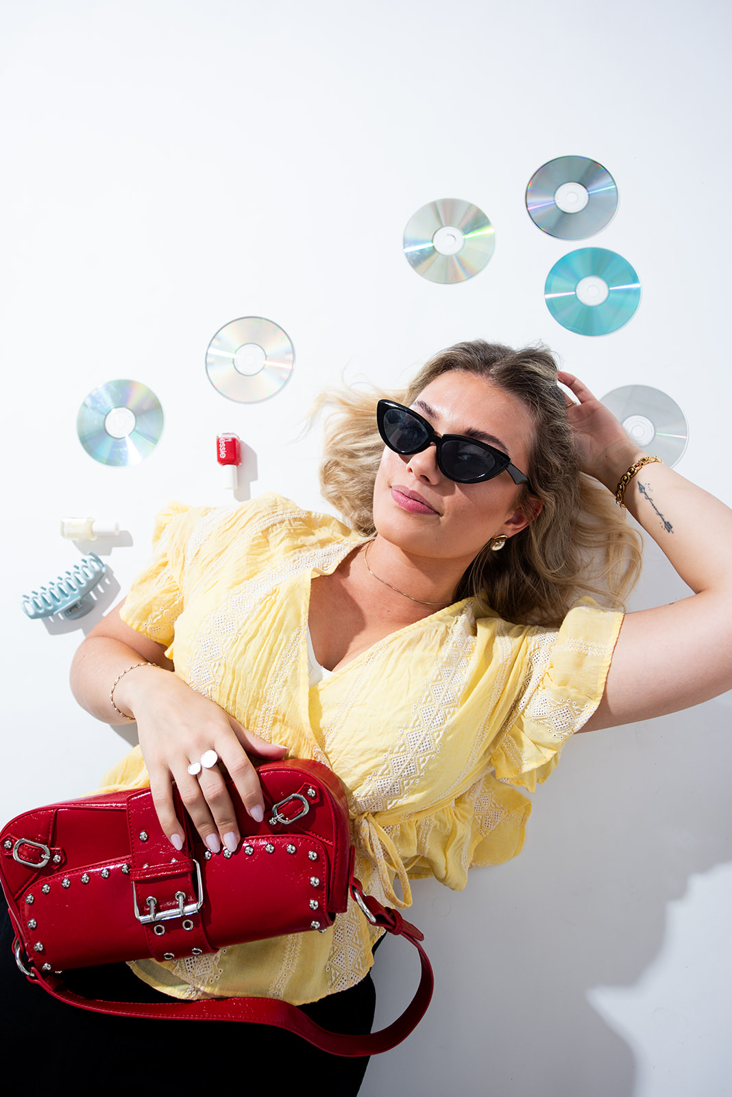Woman wearing black sunglasses lying on a studio floor surrounded by CDs and retro props during a stylized lifestyle branding photoshoot.
