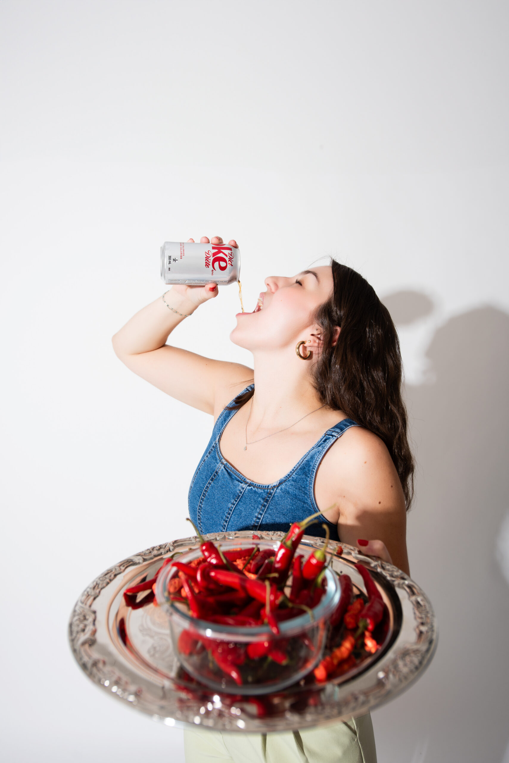 Woman in a denim tank top drinking from a Diet Coke can while holding a silver tray with red accessories, set against a white studio background, showcasing Pepper’s bold, playful content style for branding and social media marketing.