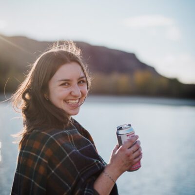 Woman smiling while holding a drink beside a lake with mountains in the background, capturing natural lifestyle imagery and candid personal branding content.