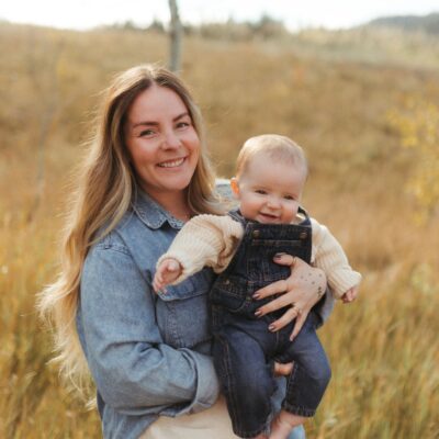 Woman smiling while holding a baby outdoors in a warm, natural field setting, dressed in neutral tones with soft golden light.