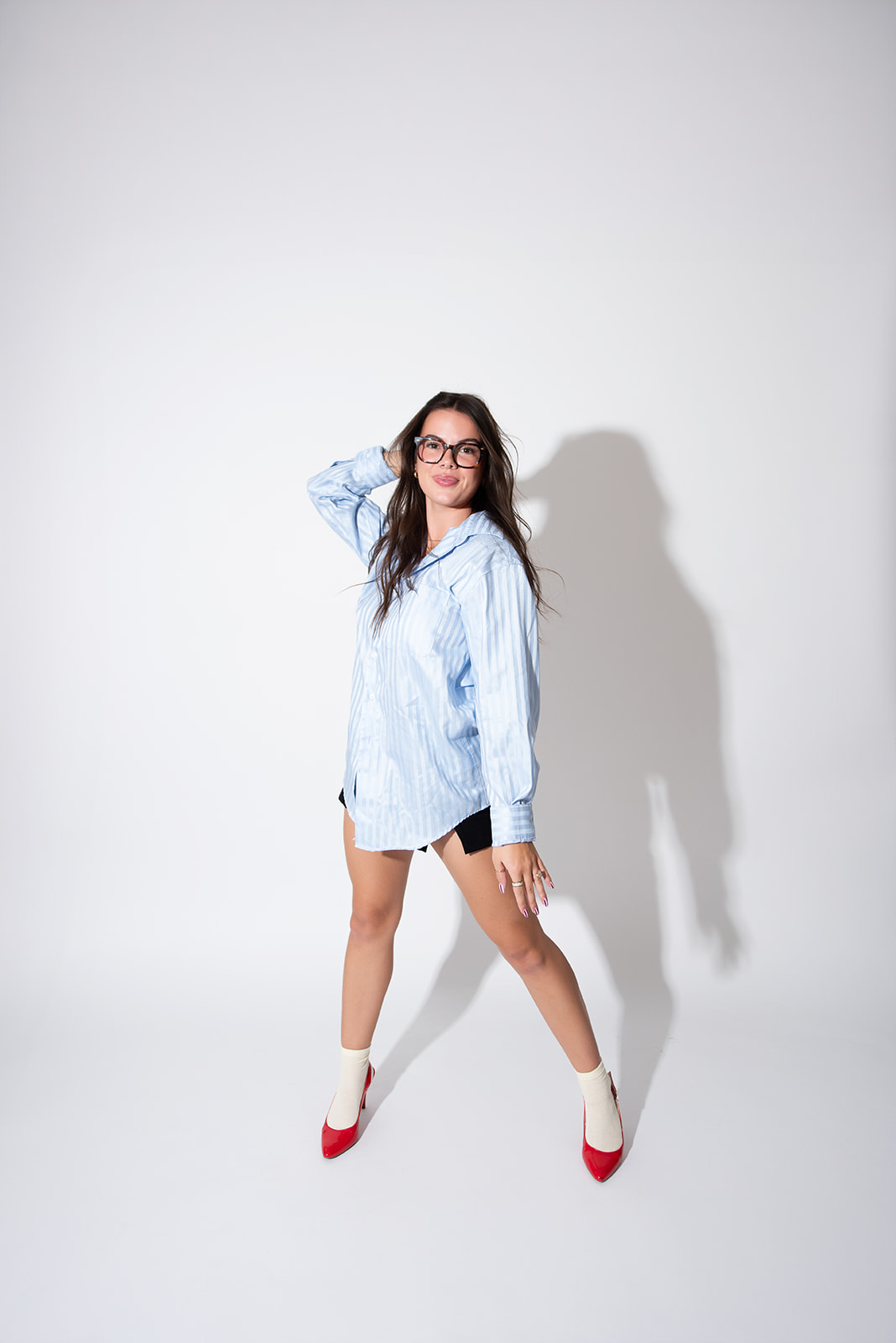 Woman in a blue striped shirt, black shorts, and red heels posing confidently in a studio with strong shadow contrast, reflecting the personality-driven content and visual identity work created by Pepper marketing agency in Kamloops.