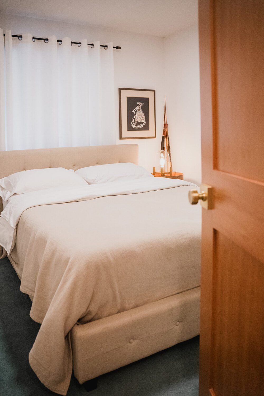 Soft neutral bedroom at The Spot with a beige upholstered bed, white curtains, warm lighting, and wood door detail.
