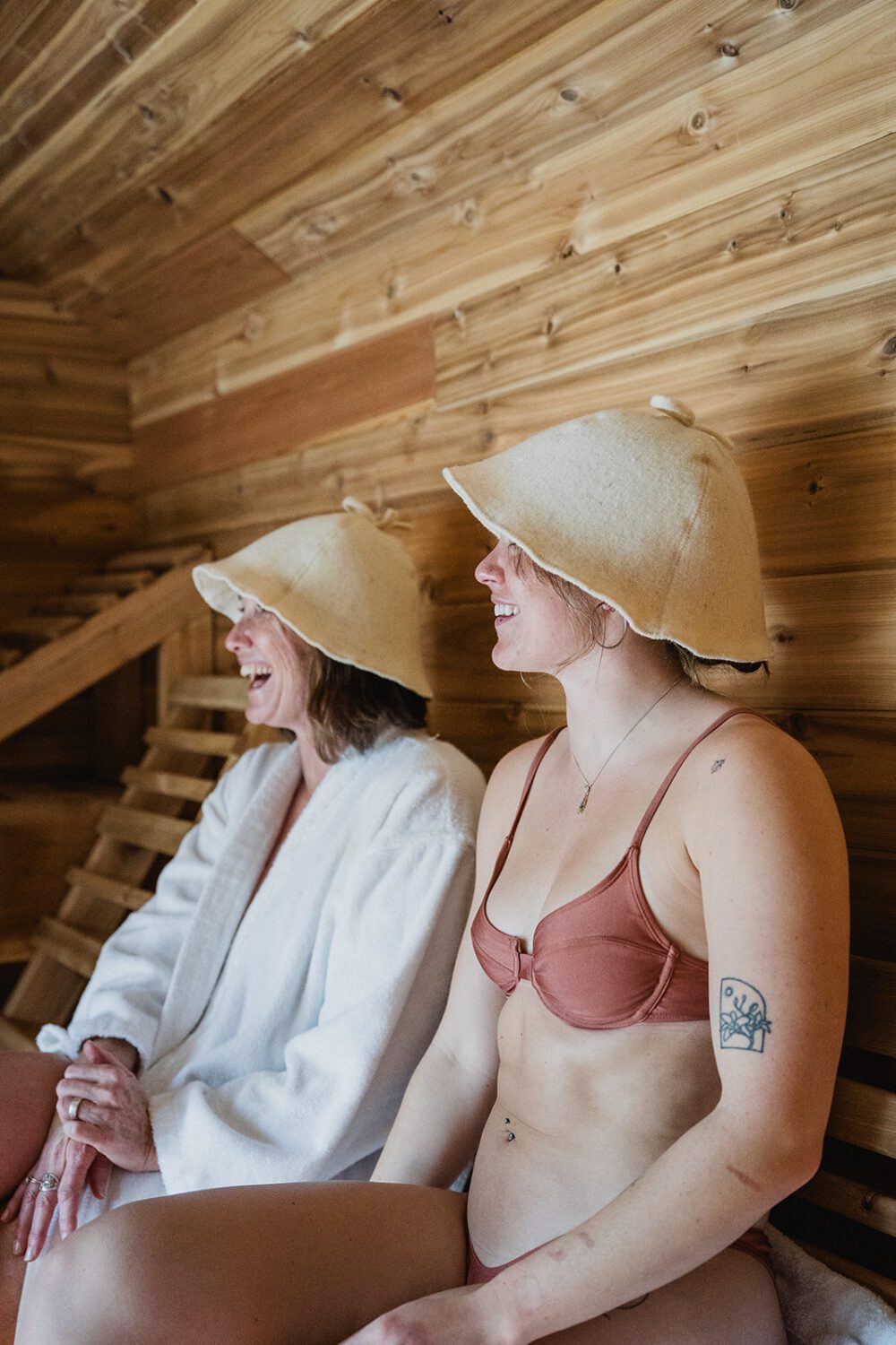 Two women relaxing and laughing inside the wood sauna at The Spot while wearing felt sauna hats.