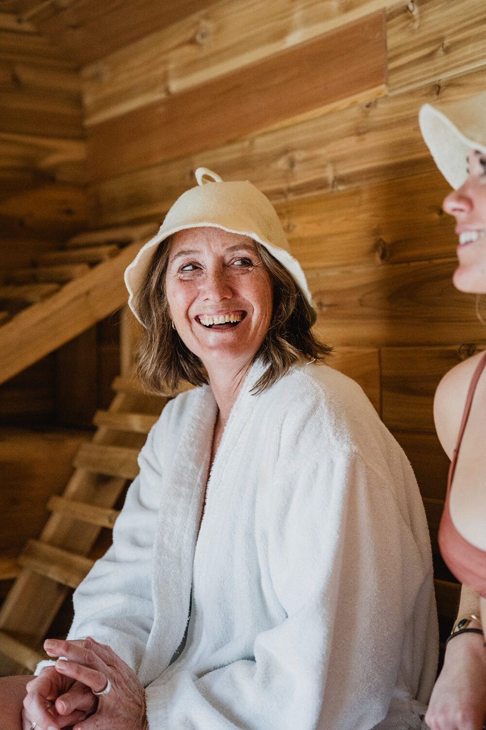 Close-up of a woman smiling inside the sauna at The Spot, wearing a white robe and felt sauna hat.