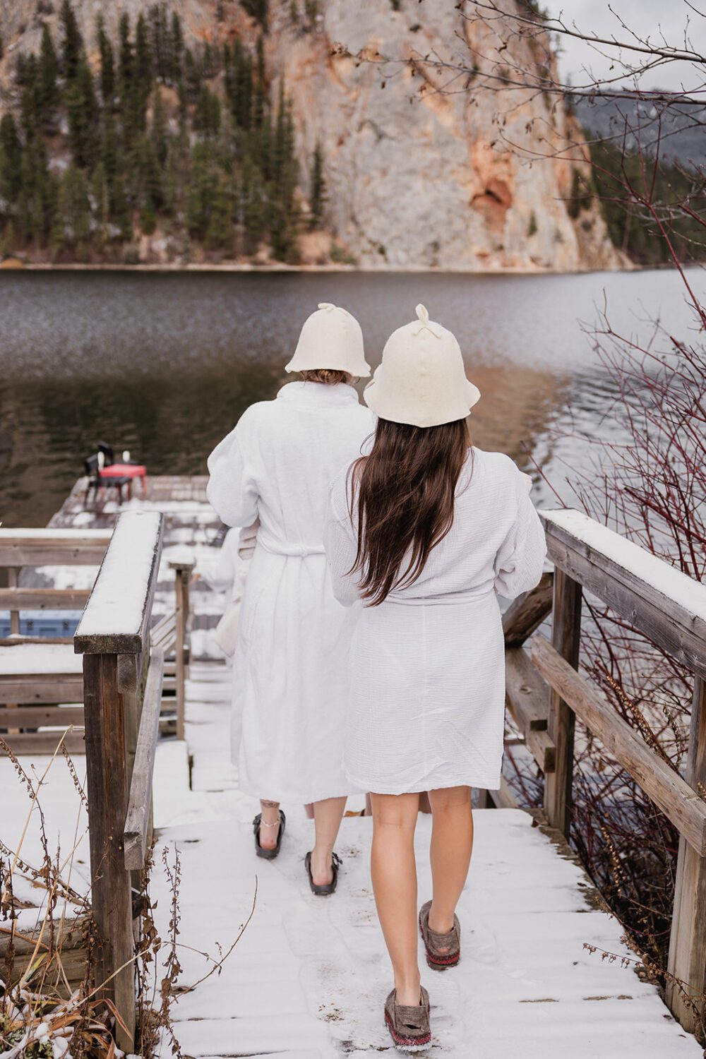 Two women in white robes and sauna hats walking down snowy stairs toward Paul Lake at The Spot.